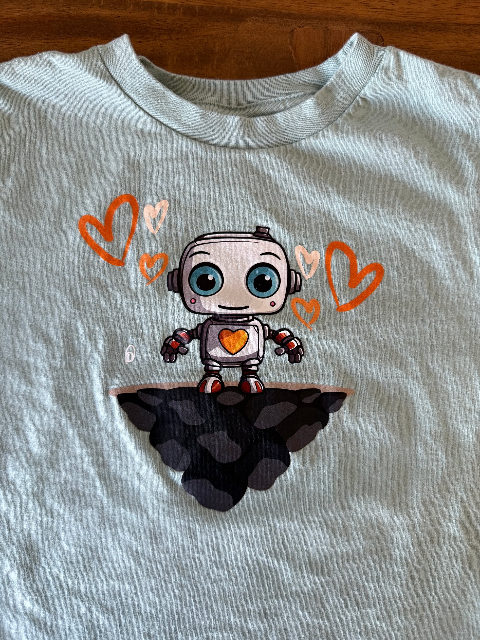 A dusty blue crewneck t-shirt laid flat on a wooden surface, featuring a cartoon robot graphic on the chest surrounded by orange hearts and a dark gray cloud.