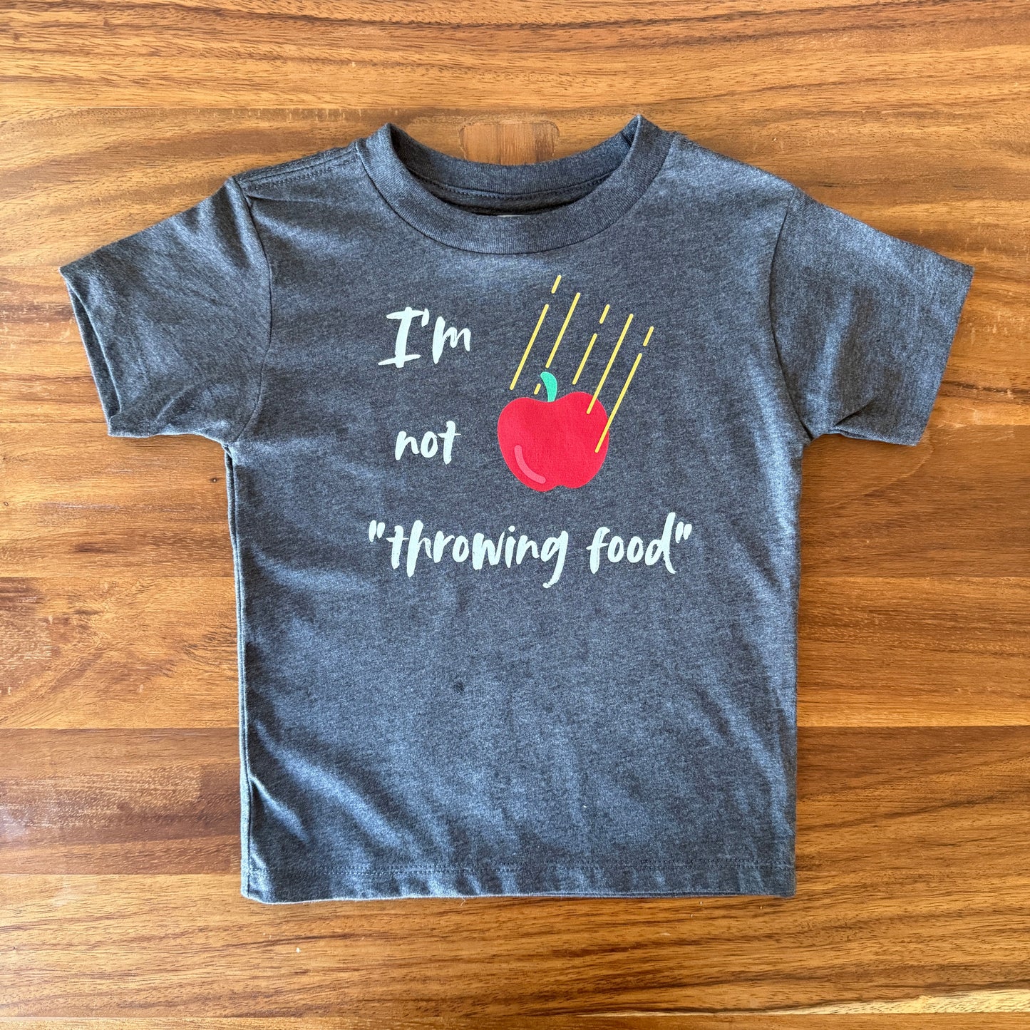 A dark heather gray short-sleeve t-shirt laid flat on a wooden table, featuring a graphic print of a red apple with yellow sticks and the text 'I'm not "throwing food"'.