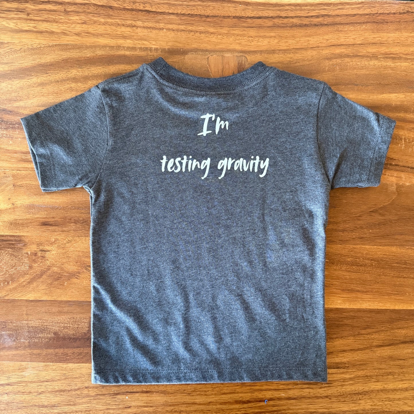 A dark heather gray t-shirt with the phrase 'I'm testing gravity' printed in white script across the back, laid flat on a wooden table.