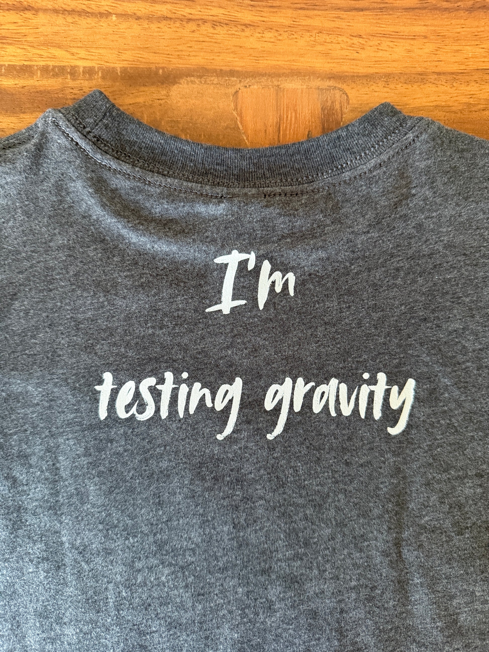 A close-up of a dark heather gray t-shirt laid flat on a wooden surface, featuring the white text 'I'm testing gravity' printed on the front.