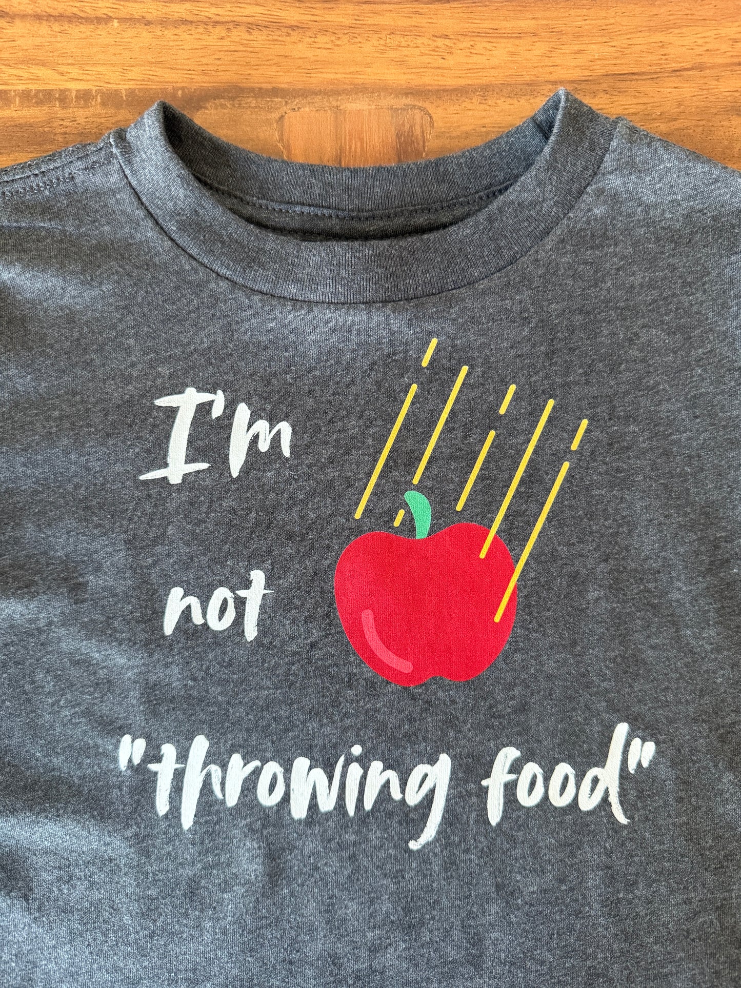 A close-up of a dark heather gray t-shirt laid flat on a wooden surface. The shirt features a graphic with the text 'I'm not throwing food' in white script, accompanied by a red apple with a green stem and yellow diagonal lines.