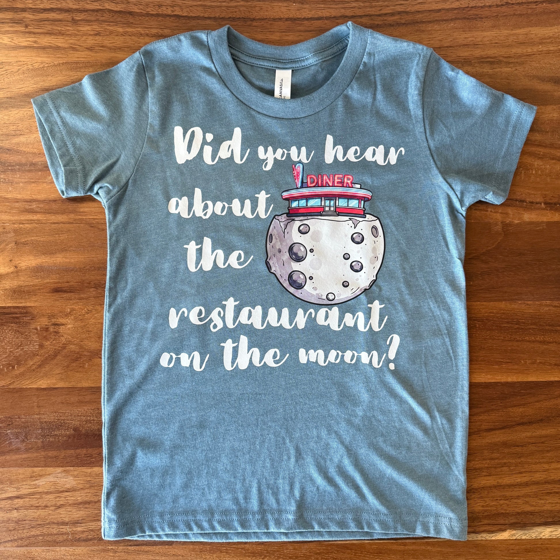 A heathered blue short-sleeve t-shirt laid flat on a wooden table, featuring a graphic print of a moon with a diner on top and the text 'Did you hear about the restaurant on the moon?'.