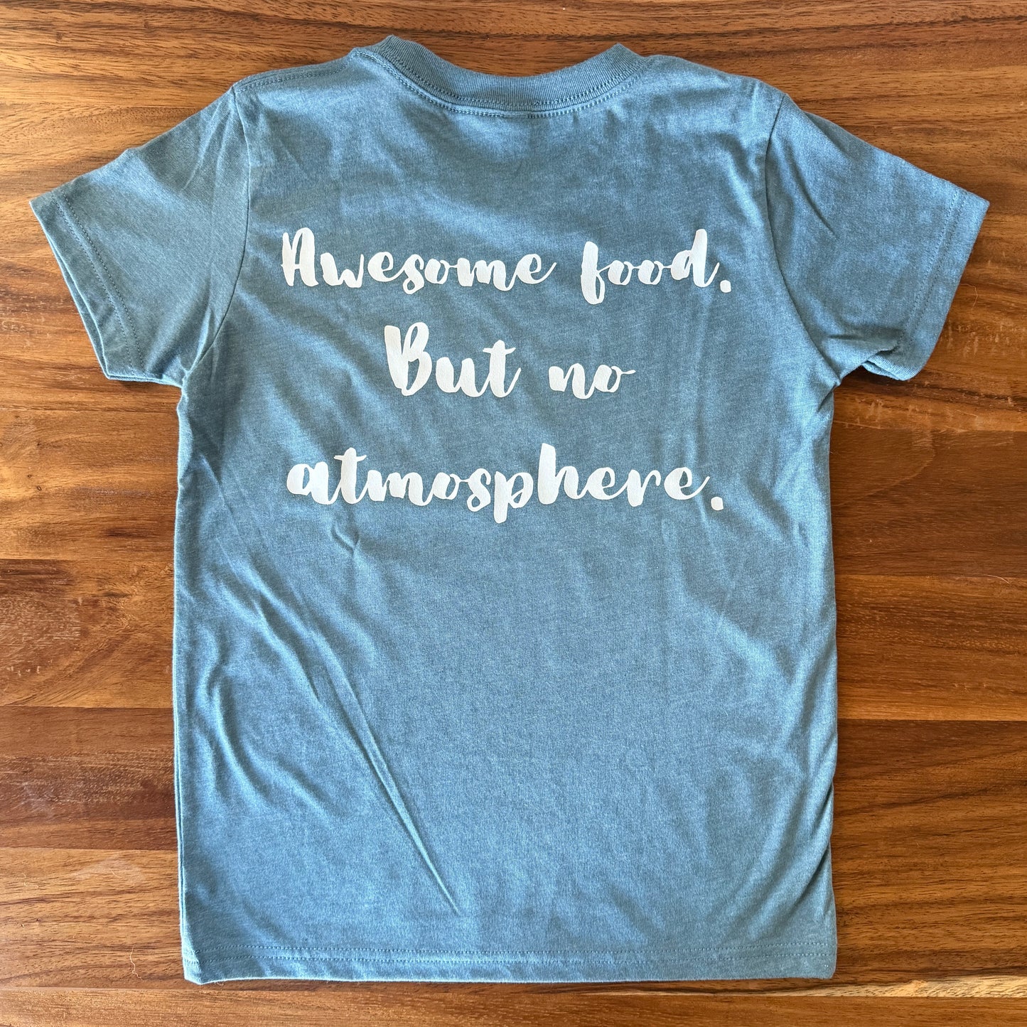 A heathered blue t-shirt with white cursive text reading 'Awesome food. But no atmosphere.' laid flat on a wooden table.