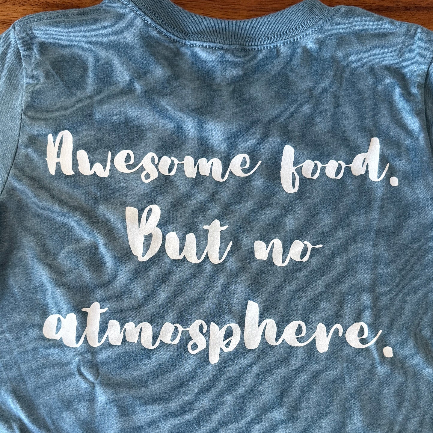 A heather blue short-sleeve t-shirt laid flat on a wooden surface, featuring the phrase 'Awesome food. But no atmosphere.