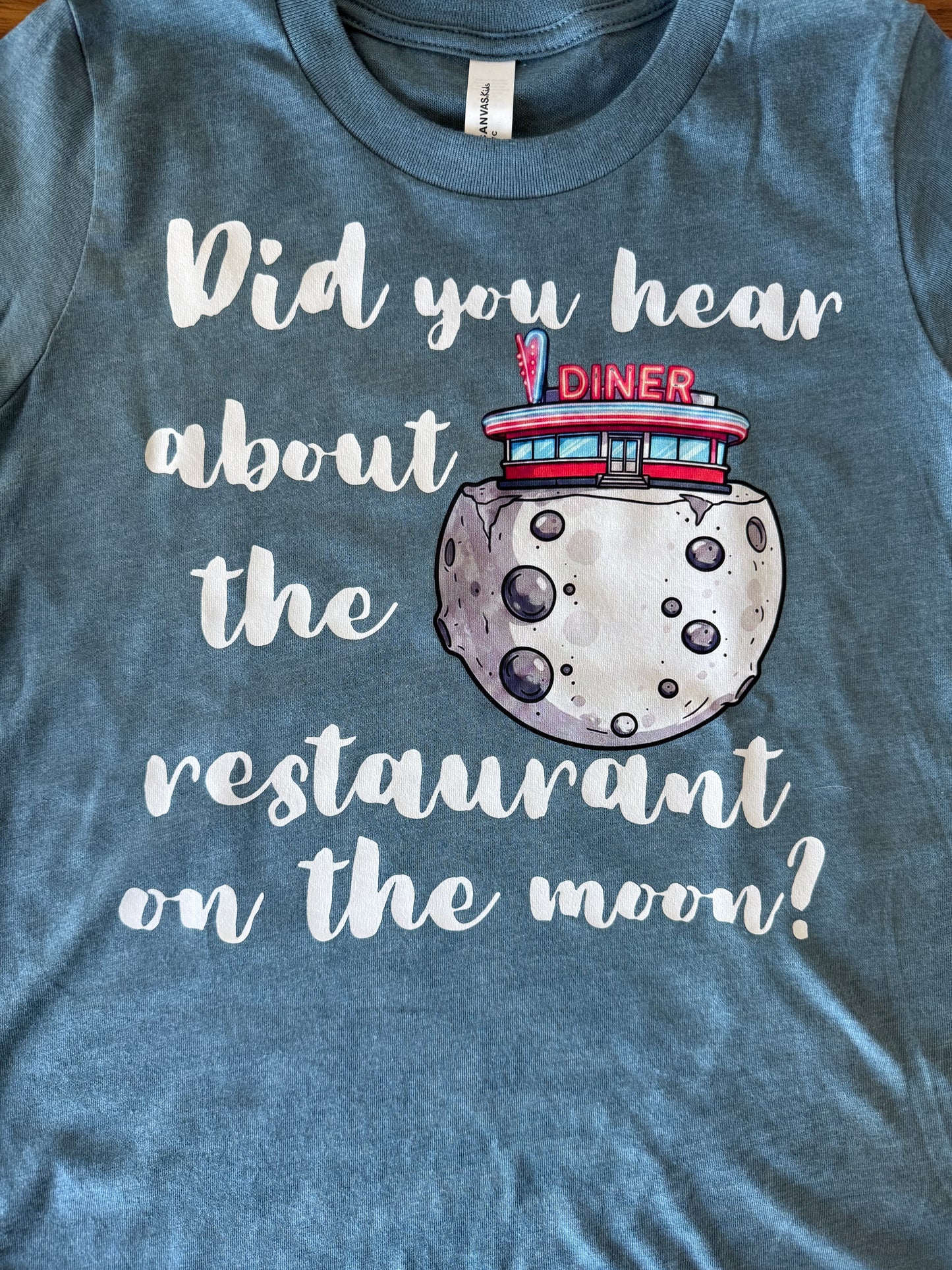 A heathered slate blue t-shirt featuring a graphic print of a moon with a red and blue diner on top, accompanied by the white text 'Did you hear about the restaurant on the moon?'.
