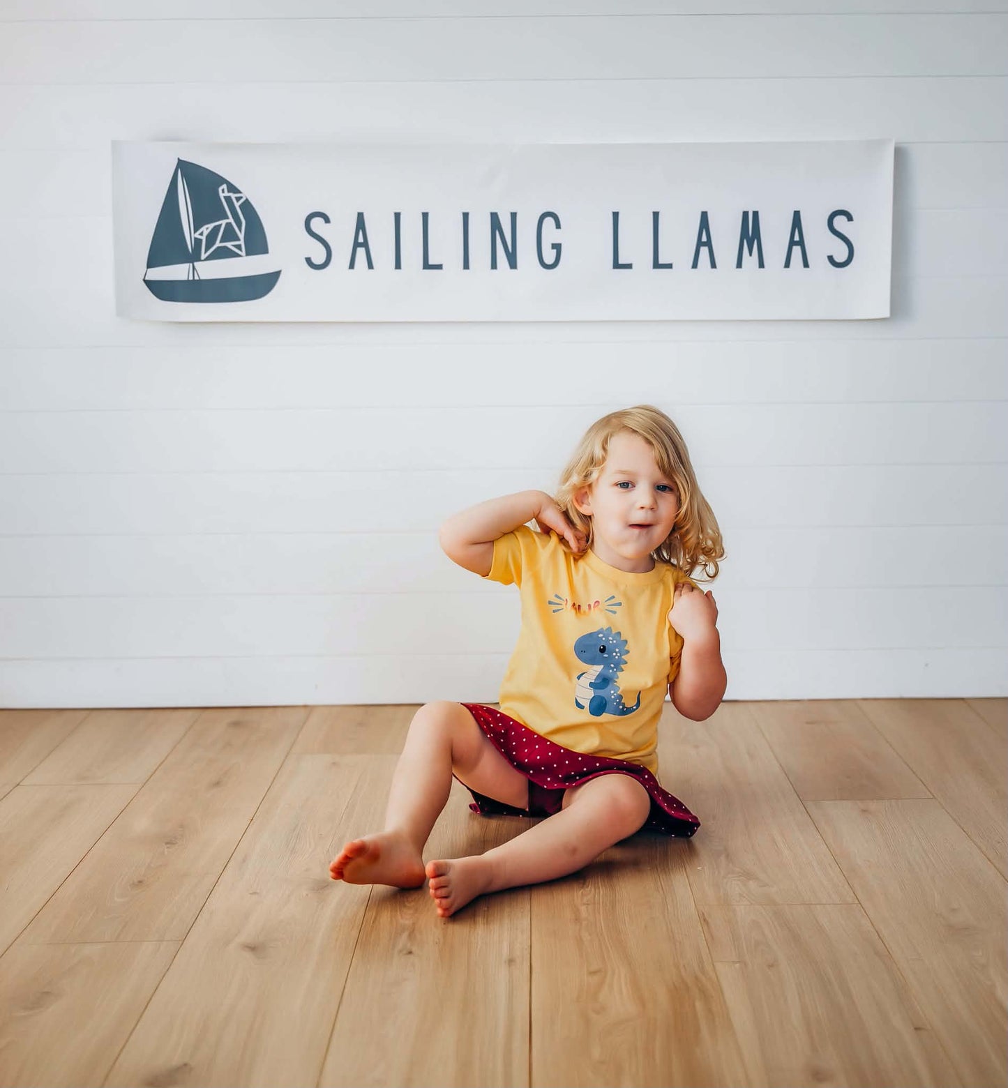 A young child sits on a light wood floor in front of a white wall with a large 'SAILING LLAMAS' banner. The child is wearing a yellow t-shirt with a blue llama graphic and a red patterned skirt.