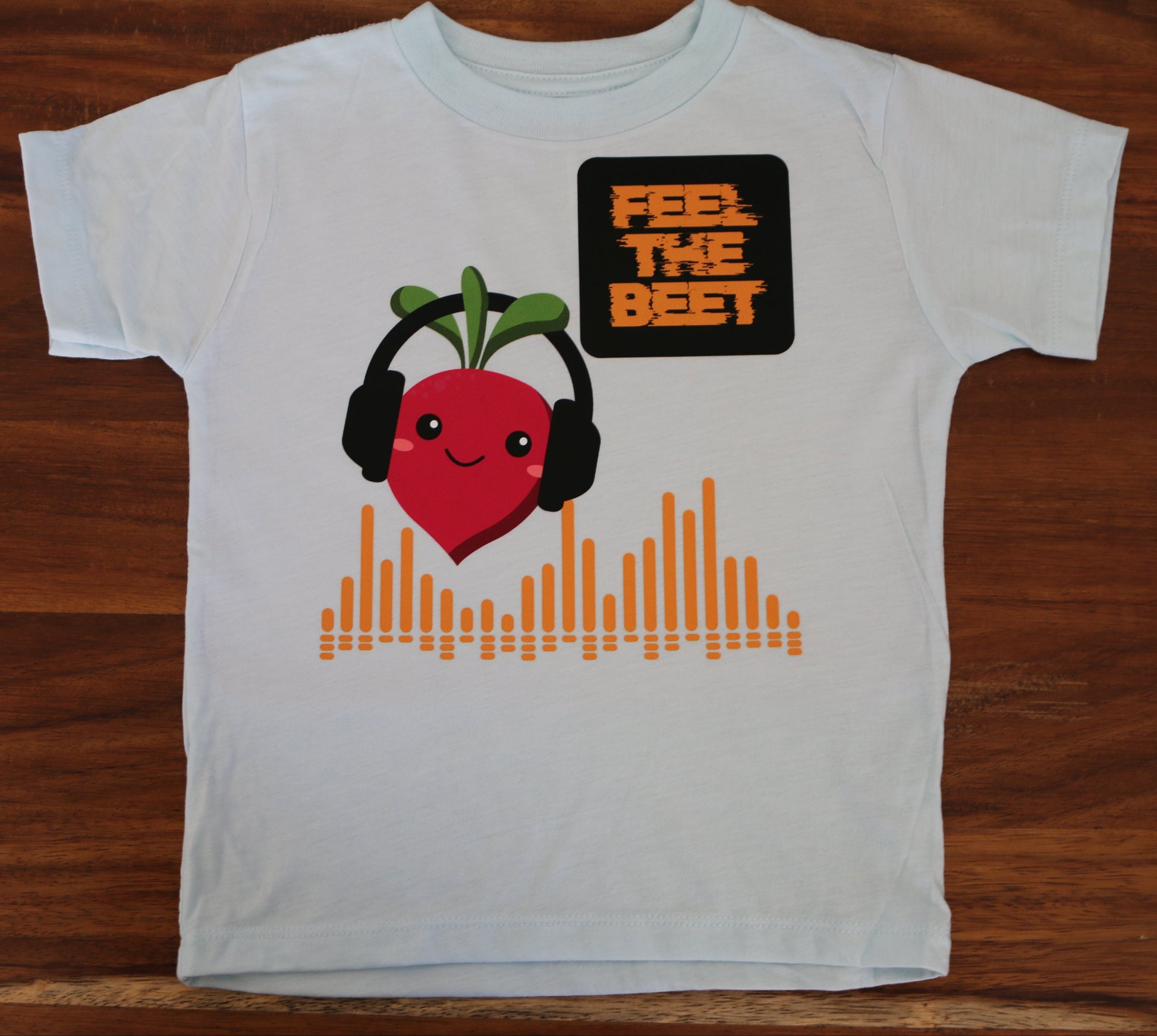 White t-shirt with a cartoon beet wearing headphones and sound waves on a wooden surface
