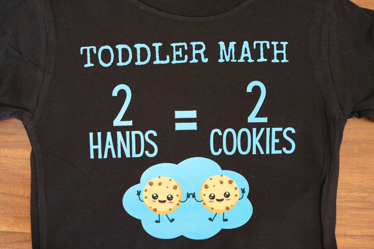 Black t-shirt with 'TODDLER MATH 2 HANDS = 2 COOKIES' text and cookie graphics on a wooden surface.