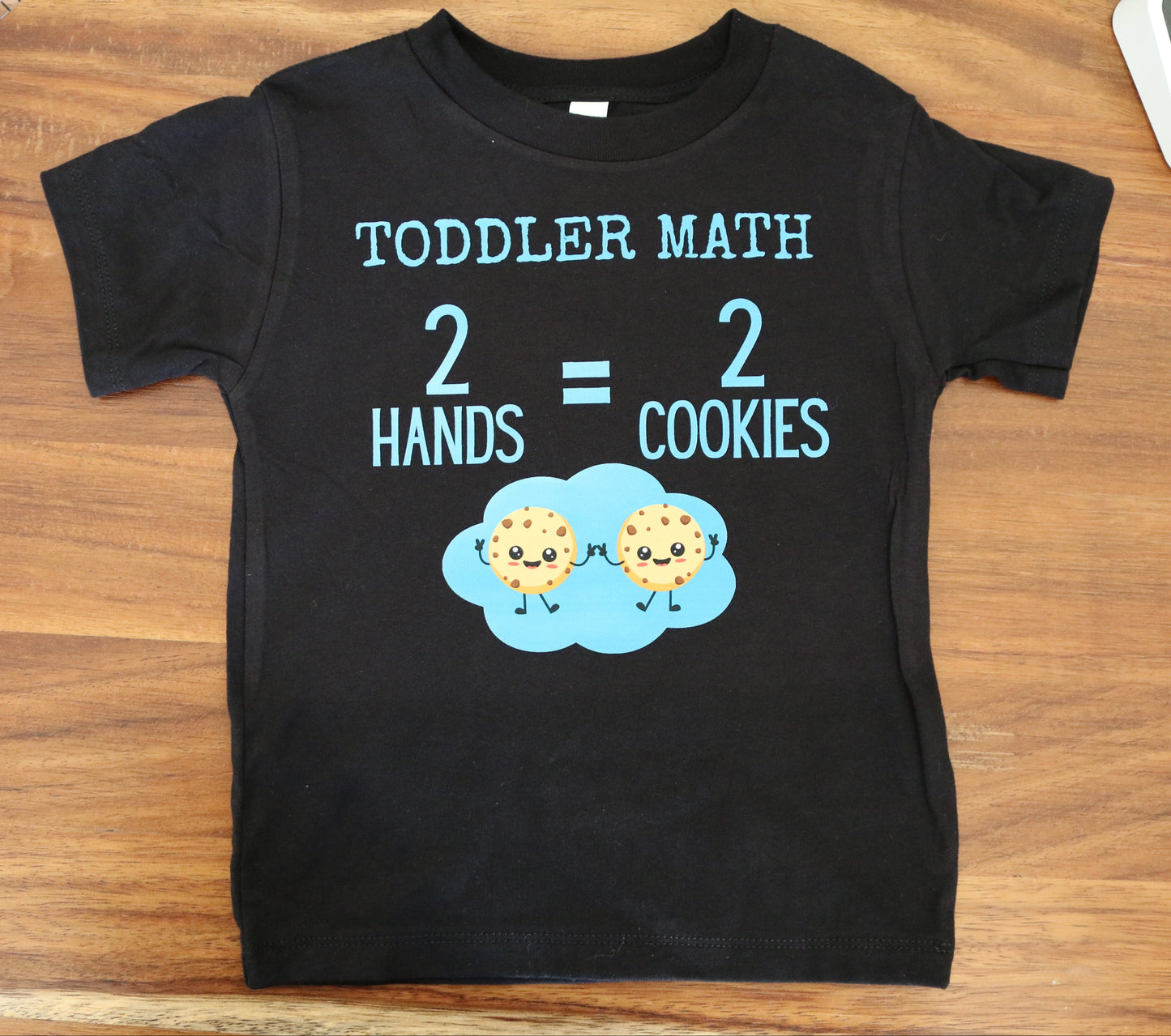 Black t-shirt with 'TODDLER MATH' text and cookie graphics on a wooden surface