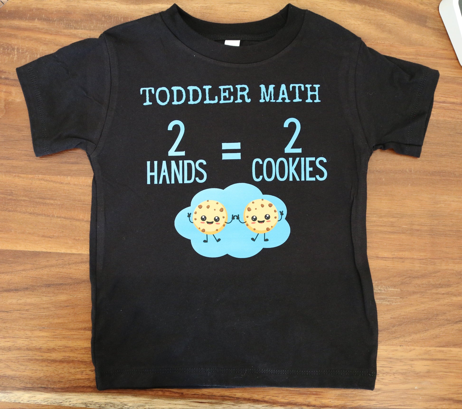 Black t-shirt with 'TODDLER MATH' text and cookie graphics on a wooden surface